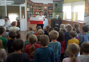Stojące dzieci, prowadząca trzyma flagę Polski. Mężczyzna gra na pianinie.